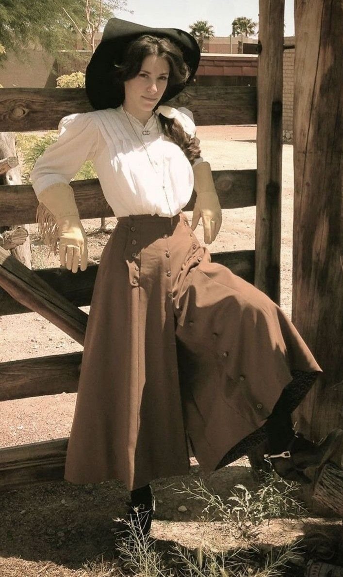 Woman in 19th century western attire posing by a rustic wooden fence.