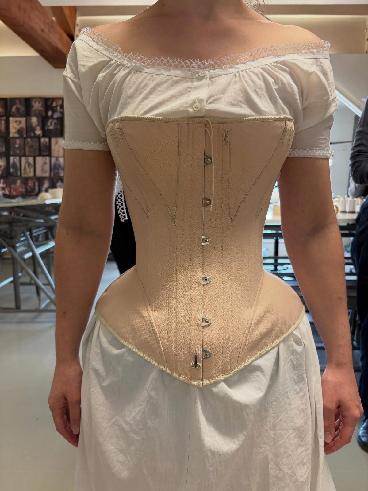 Person wearing a tan Victorian-style corset over a white off-the-shoulder chemise.