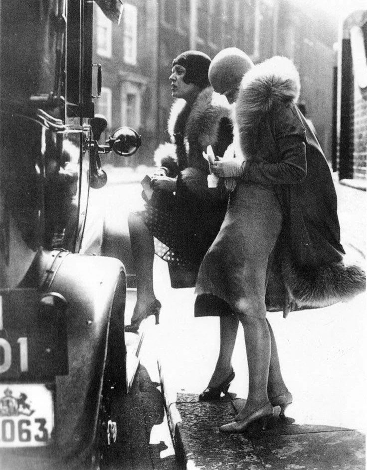 Two women in 1920s attire wearing high heels standing by a vintage car.