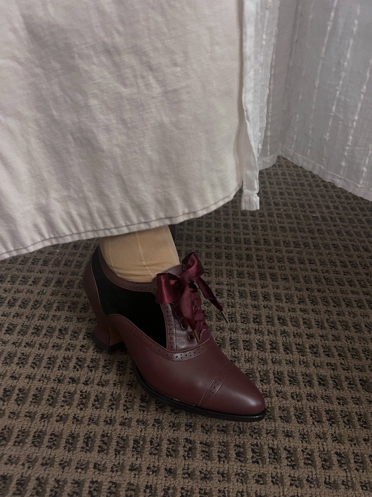 Burgundy leather lace-up heels with a bow and beige socks on carpet.