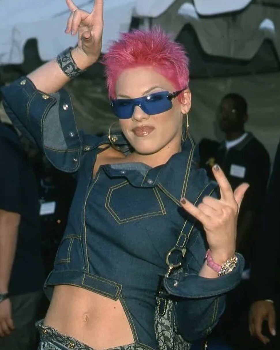 Woman with short pink hair wearing a cropped denim jacket and blue sunglasses.