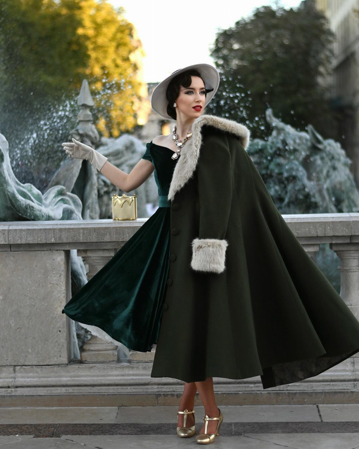 Woman in dark green 1950s velvet dress and matching fur-trimmed cape by fountain