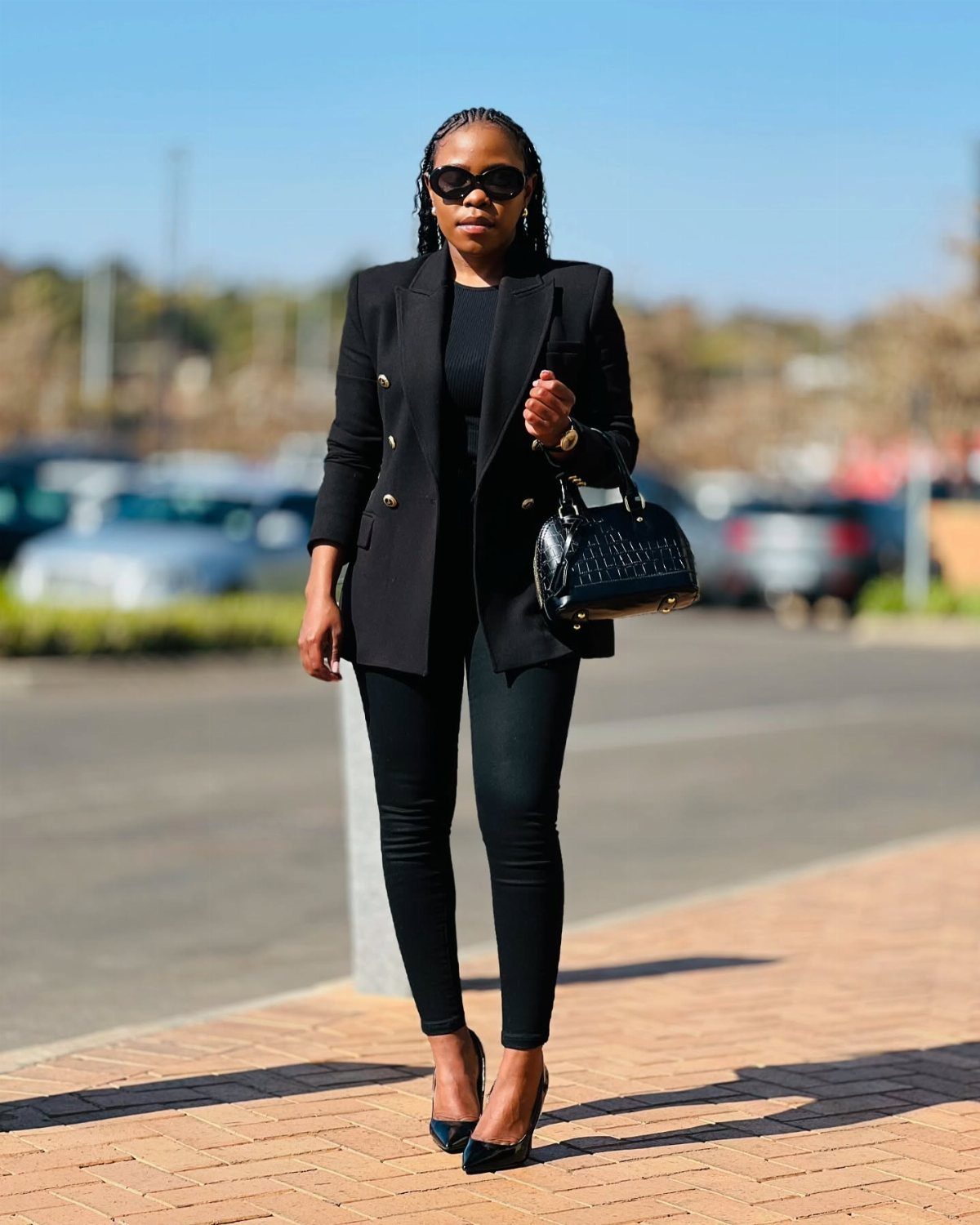 Woman in black blazer, skinny pants, and heels posing outdoors with black handbag.