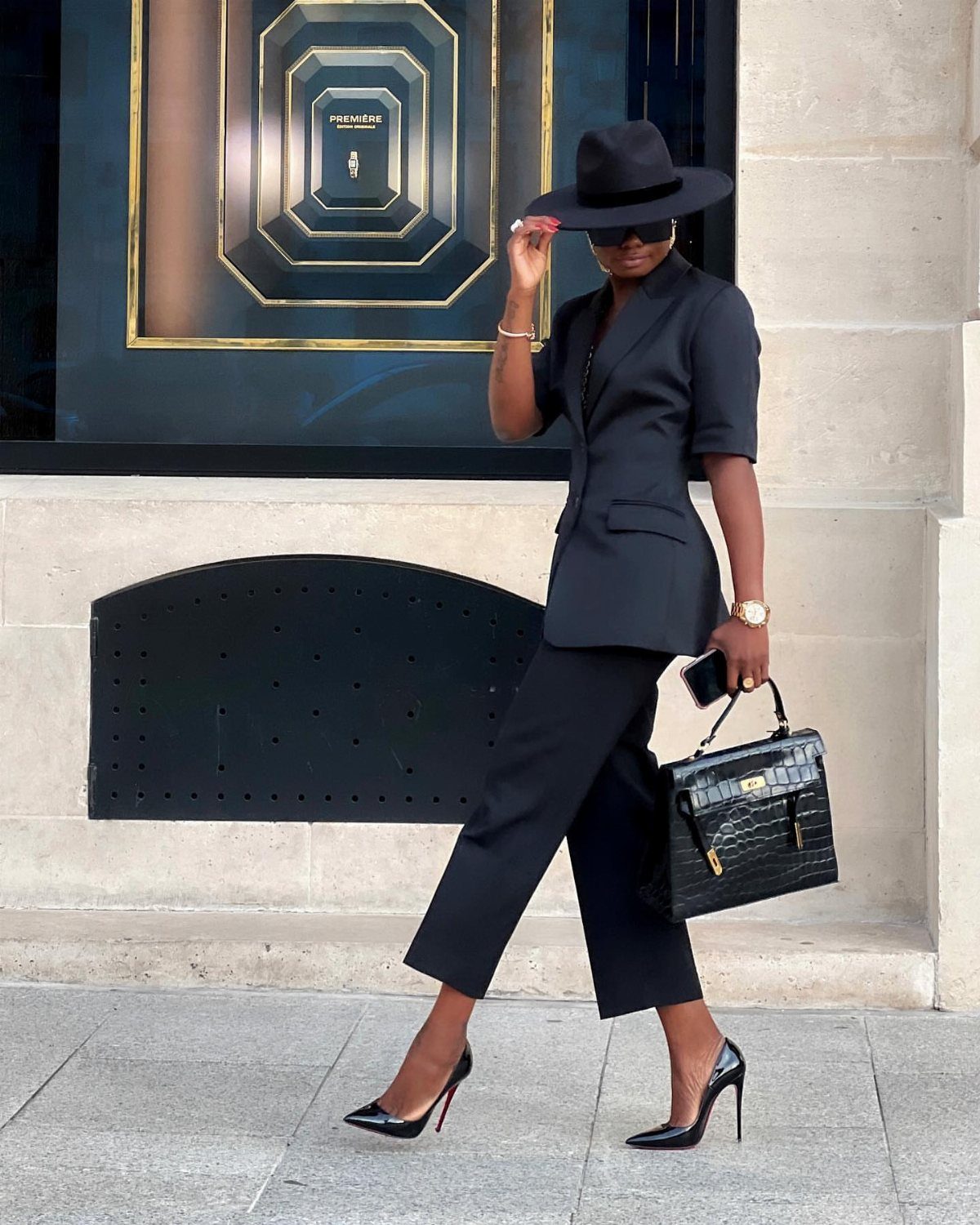 Woman in black blazer, cropped trousers, and wide-brimmed hat walking on city sidewalk.