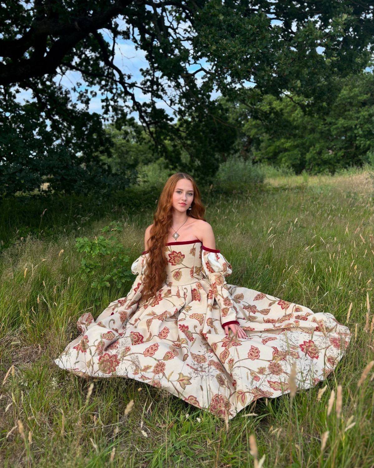 Woman in a cream and red floral Tudor-style gown sitting in a grassy field.
