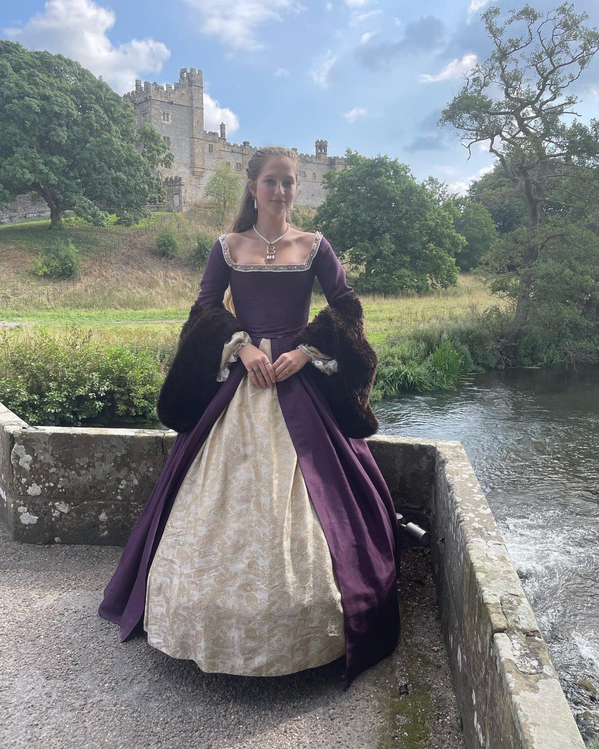 Woman in a purple and cream Tudor gown standing before a stone castle.