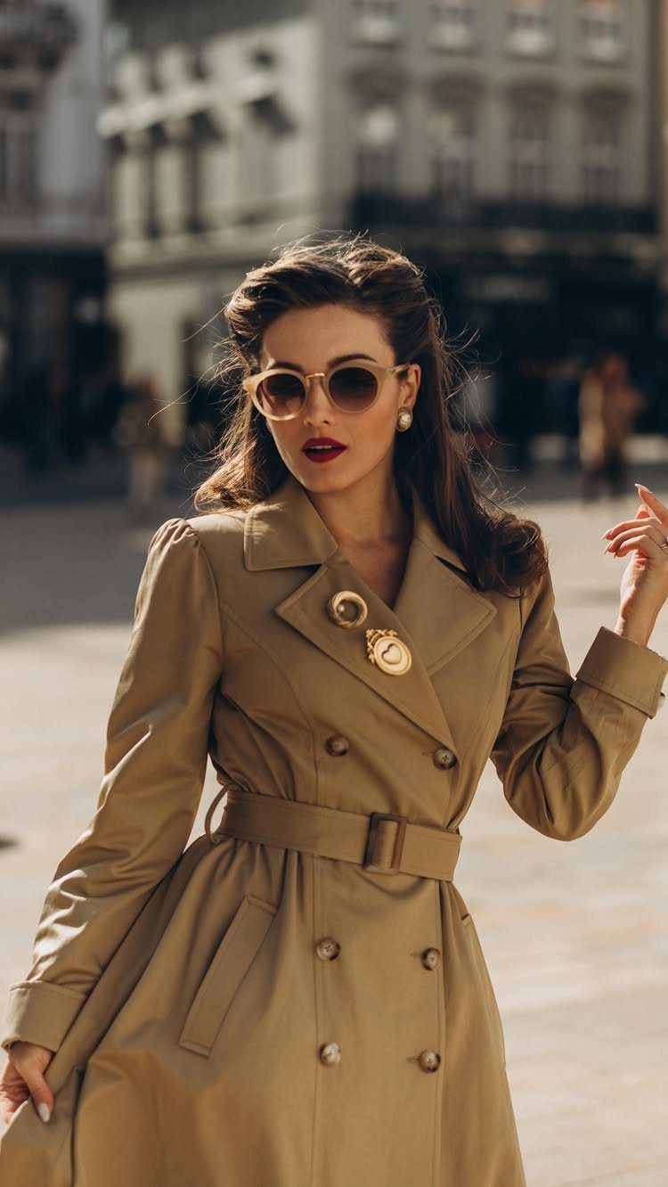 Woman wearing a tan belted trench coat and sunglasses in a city setting.