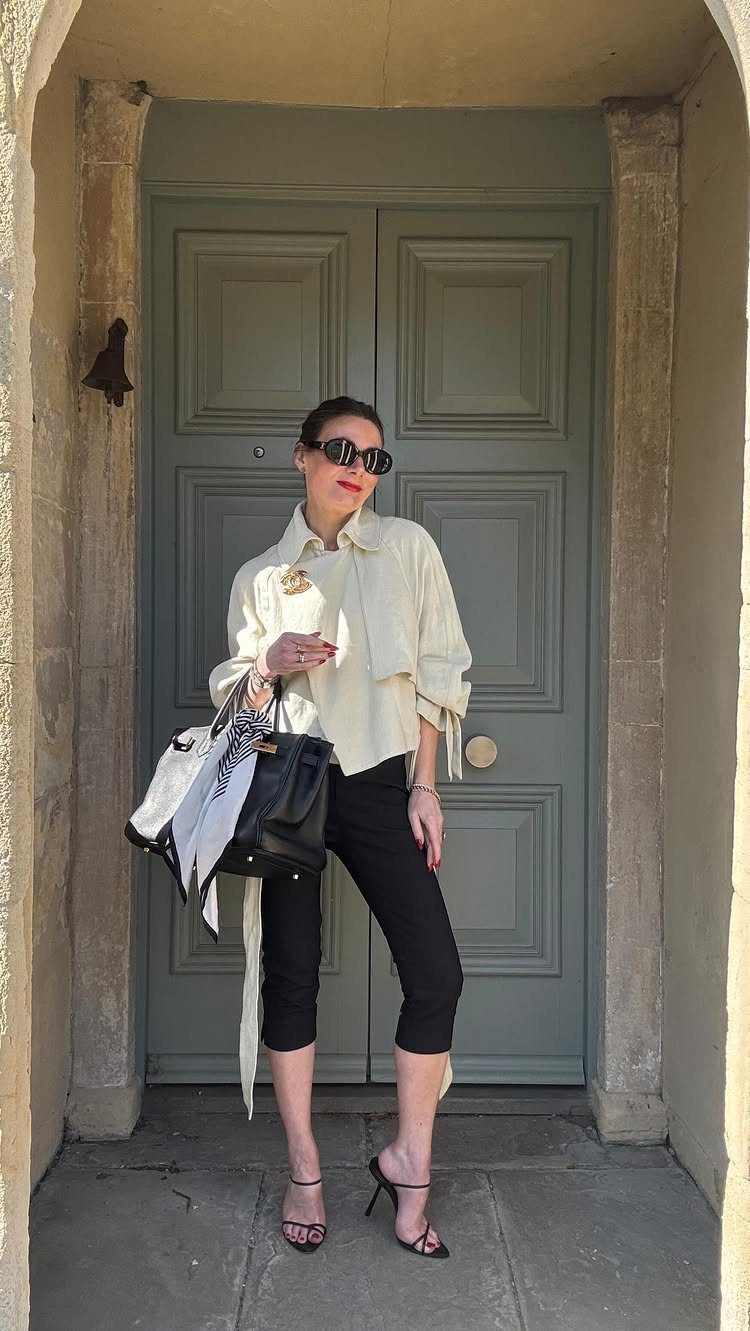 Woman in cream button-down shirt and black cropped trousers standing by grey door