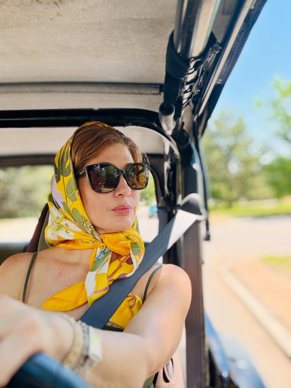 Woman in a vintage car wearing a yellow patterned headscarf and dark sunglasses.