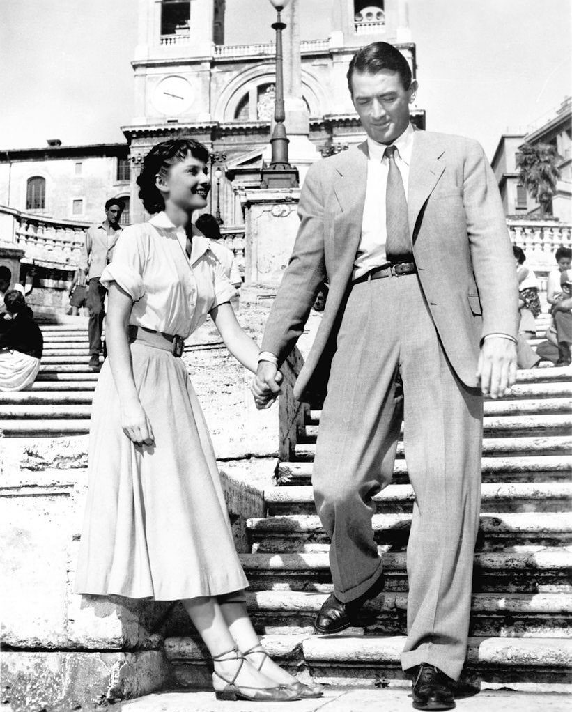 Audrey Hepburn and Gregory Peck holding hands on stone steps in Roman attire.