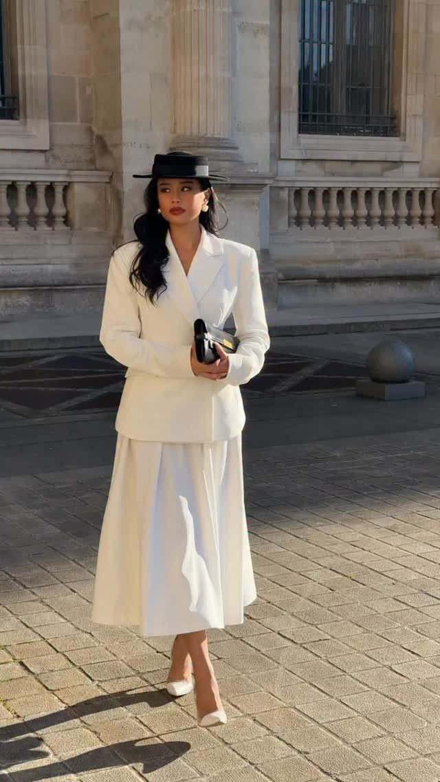 Woman in a white suit and black hat posing before a stone building.