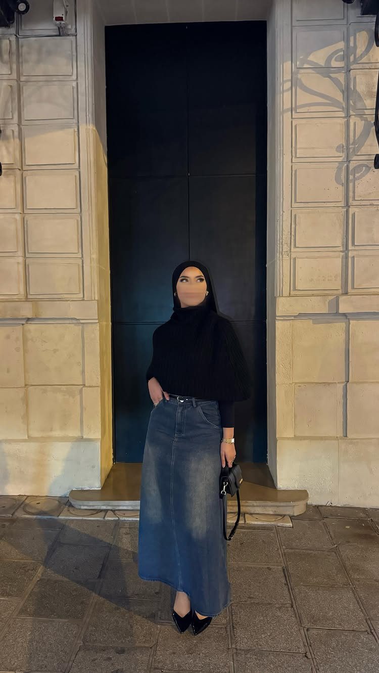 Woman in black hijab and top with a long blue denim skirt standing outdoors.