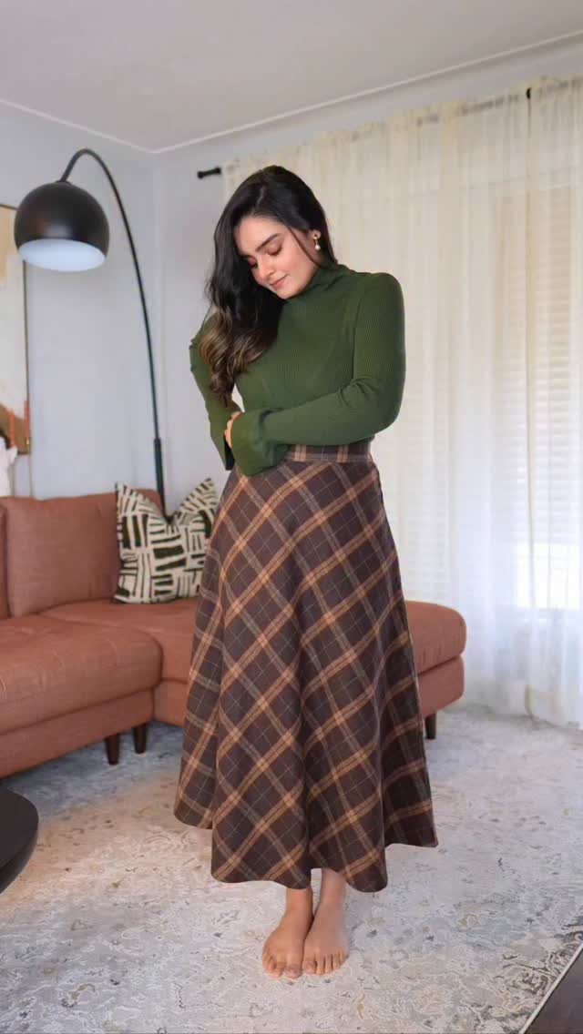 Woman wearing an olive green turtleneck and brown plaid maxi skirt indoors.