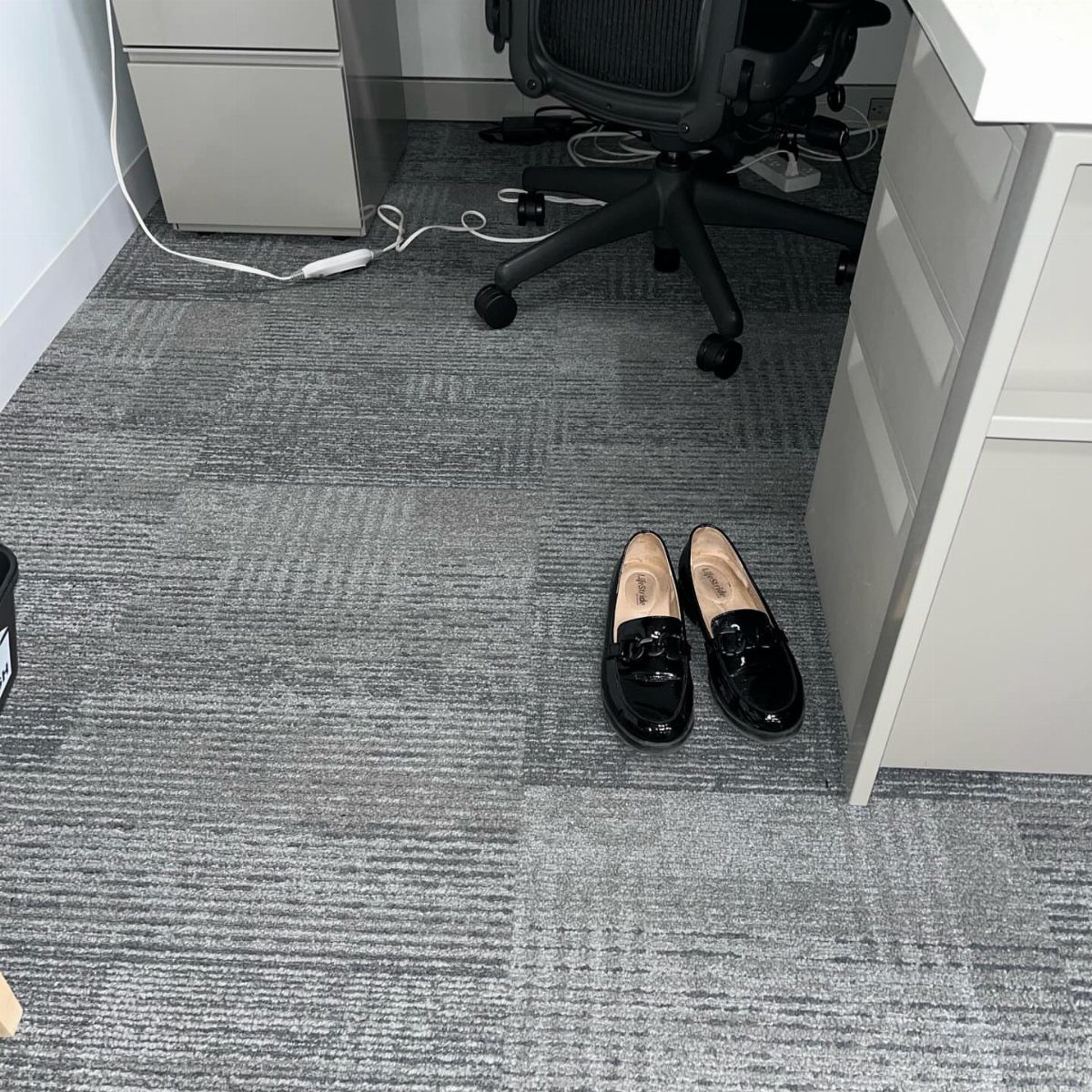 Black patent leather loafers resting on a grey office carpet near a desk.
