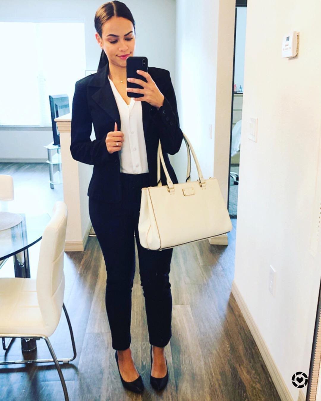 Woman in a black tailored suit and white blouse holding a white handbag.