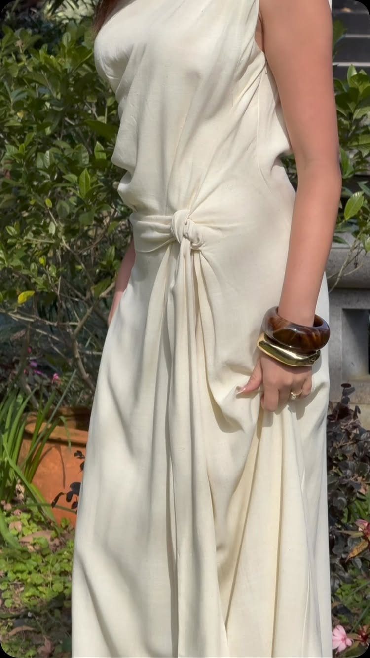 Beachy Long Dresses: 5 Ideas for Sand and Sun 3 Cream-colored long beach dress with a knot waist and chunky bracelets in a garden.