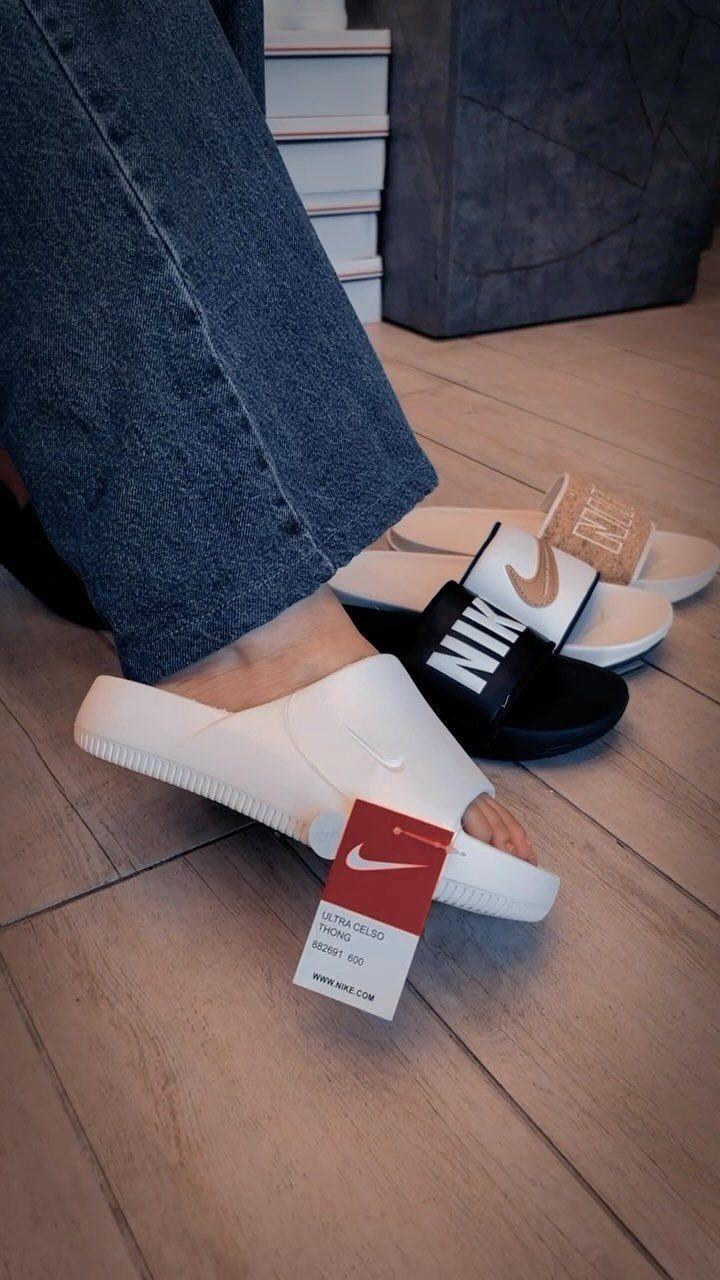 White Nike slides on a foot next to black and beige slides on wood.