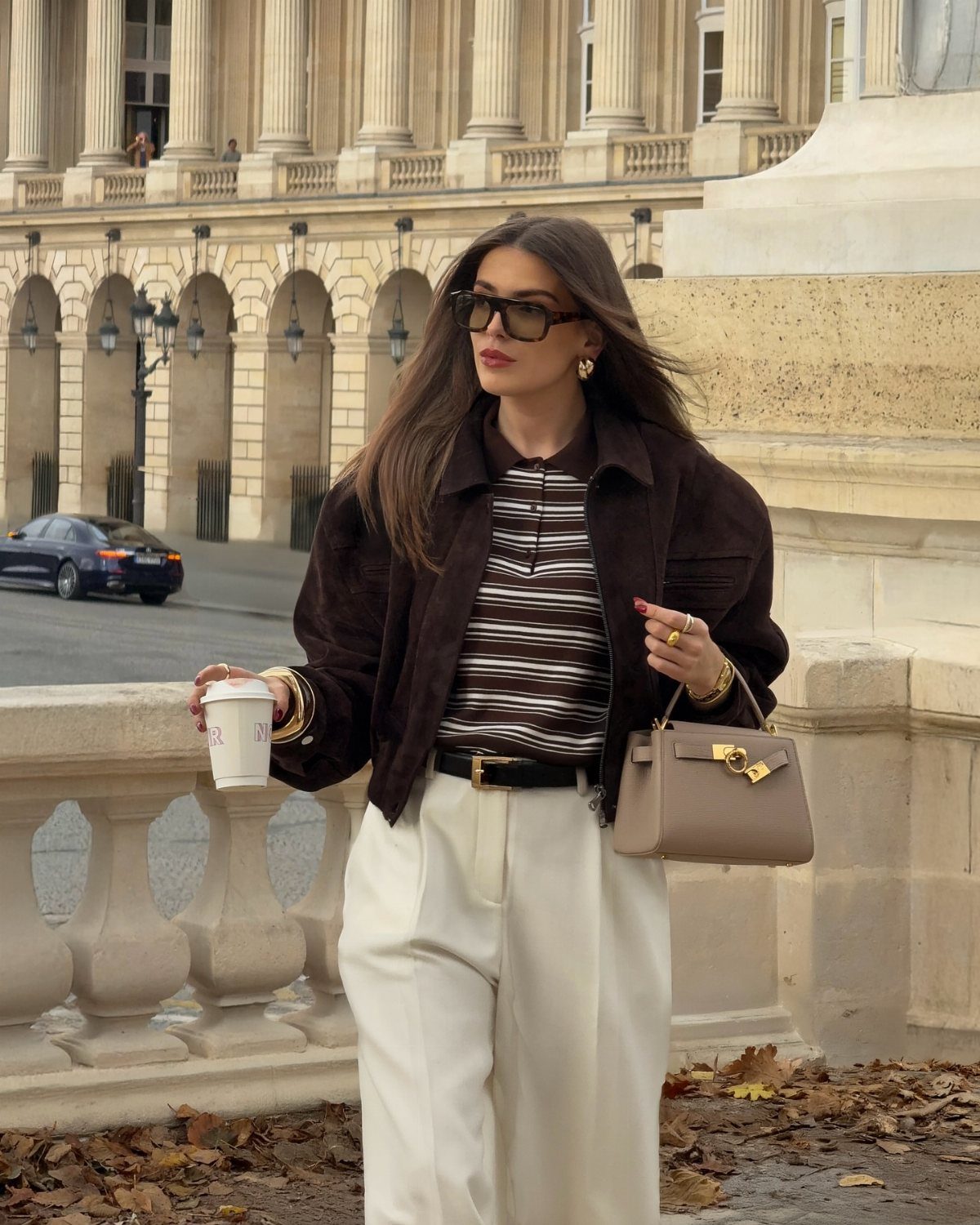 Woman wearing a brown striped polo shirt, brown jacket, and white trousers outdoors.