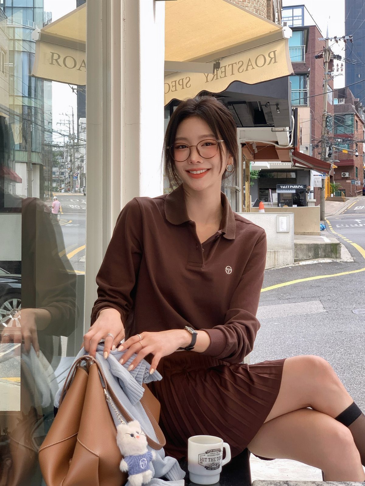 Woman in brown long-sleeved polo shirt and pleated skirt sitting outdoors with handbag.