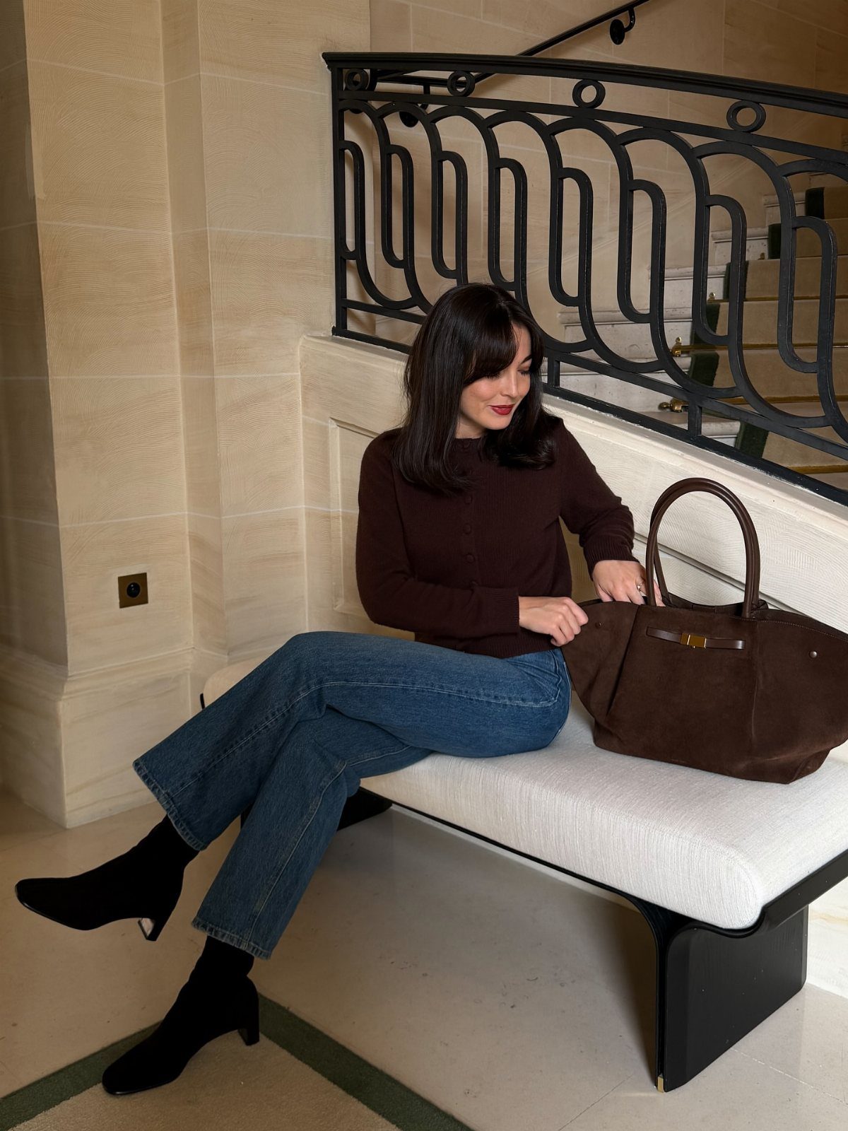 Woman in brown top and blue jeans wearing black ankle boots sitting on bench.