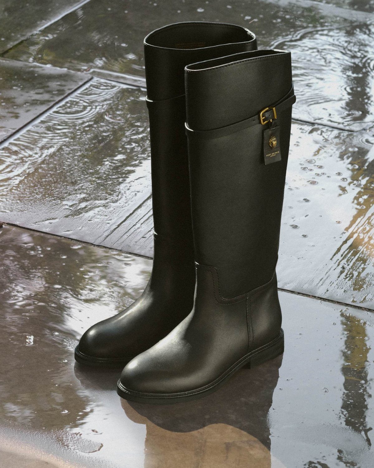 Tall black waterproof boots with gold buckles standing on wet stone pavement