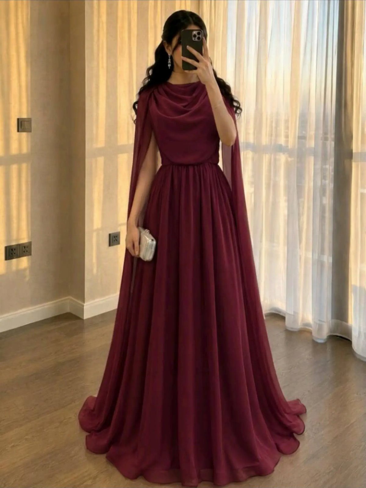 Woman in a floor-length burgundy gown with a flowing cape and silver clutch.