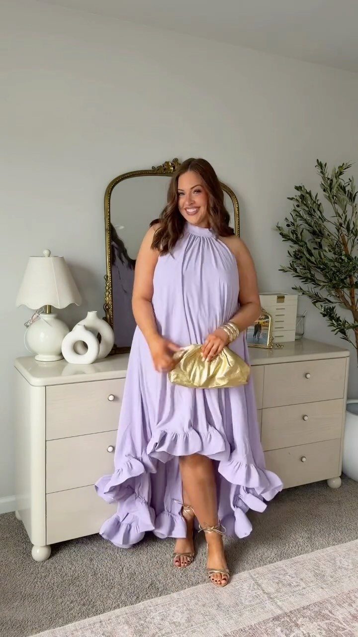 Woman in a lavender high-low dress with gold accessories standing by a dresser.
