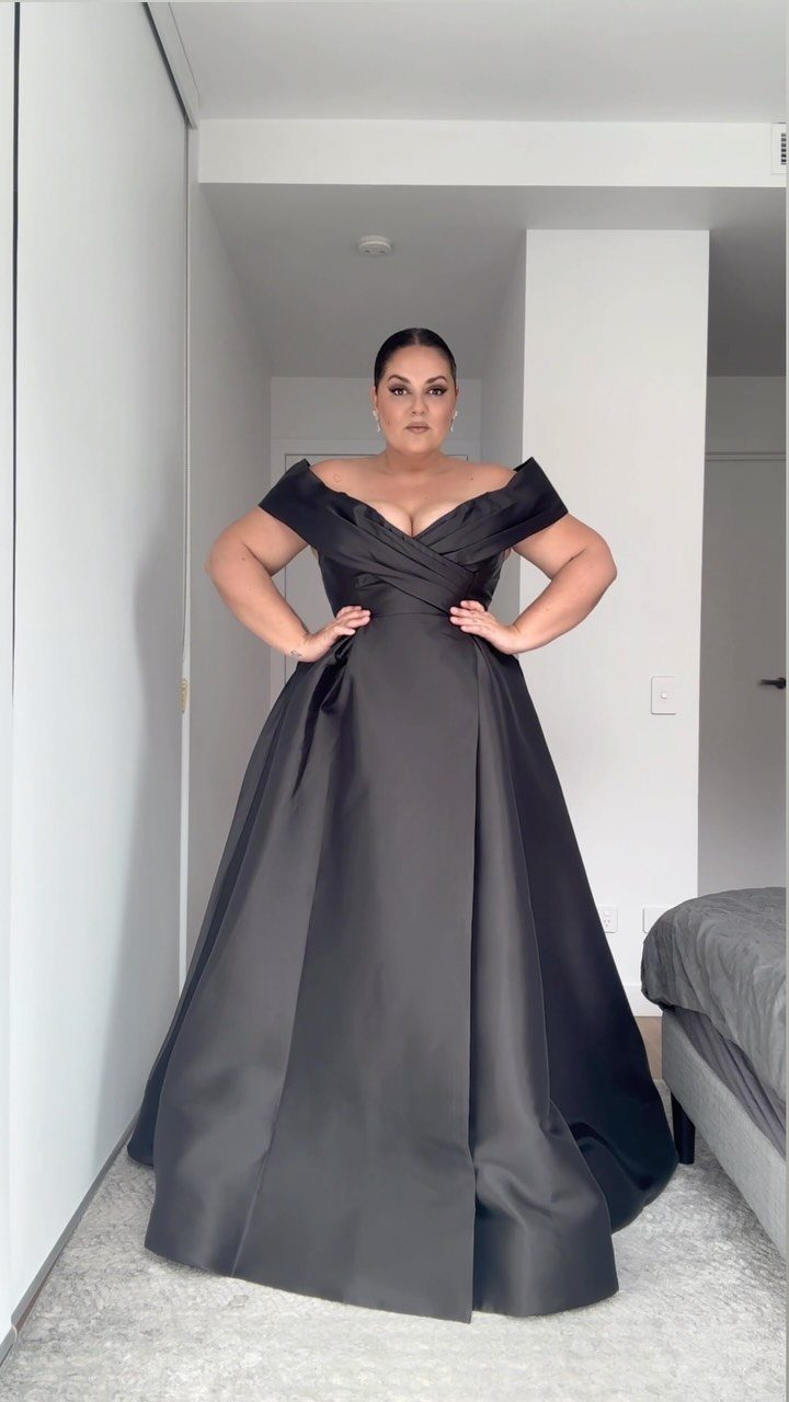 Woman wearing a voluminous off-the-shoulder black satin ball gown in a white room