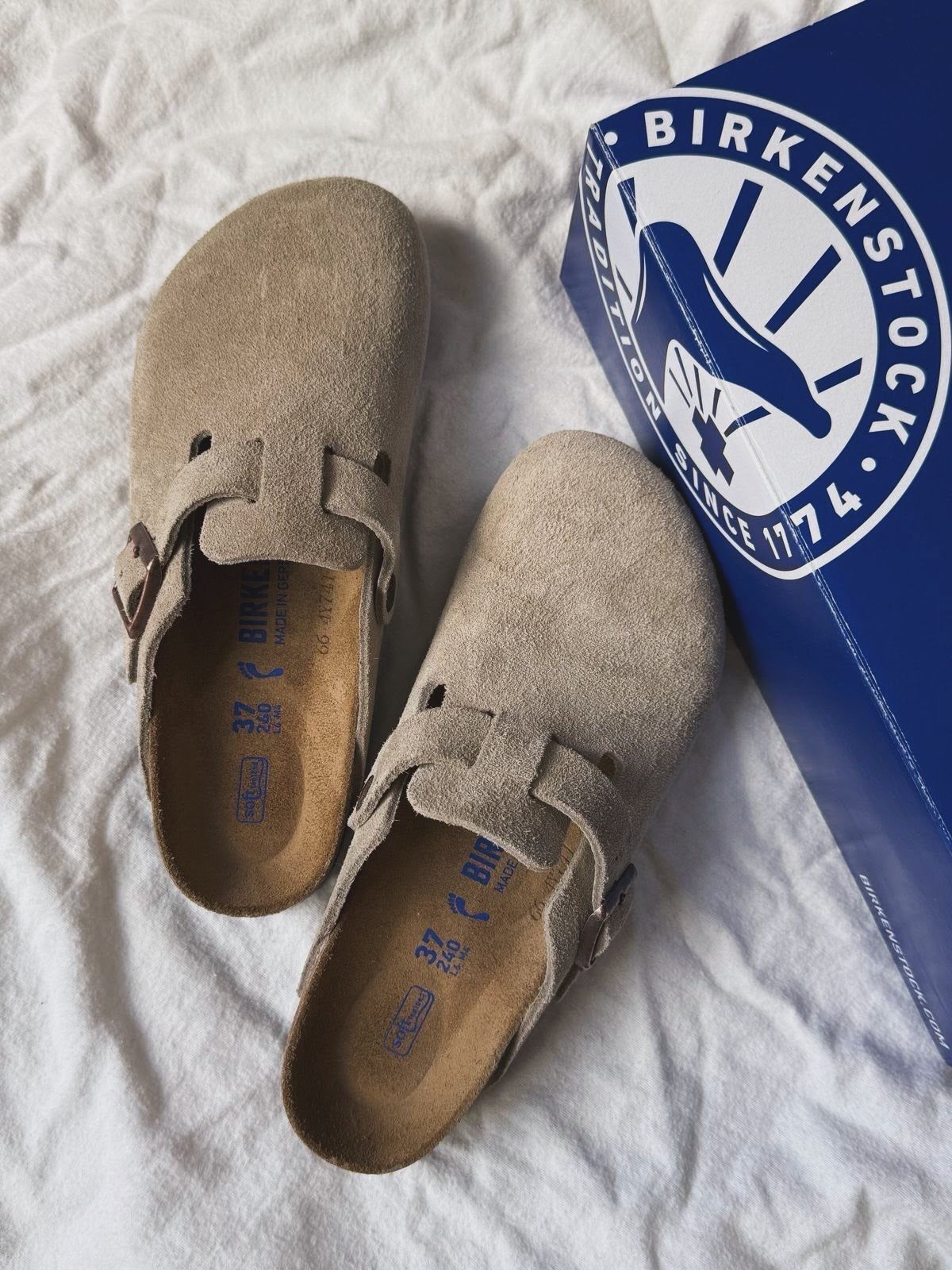 Beige suede Birkenstock clogs sitting on white fabric next to a blue box.