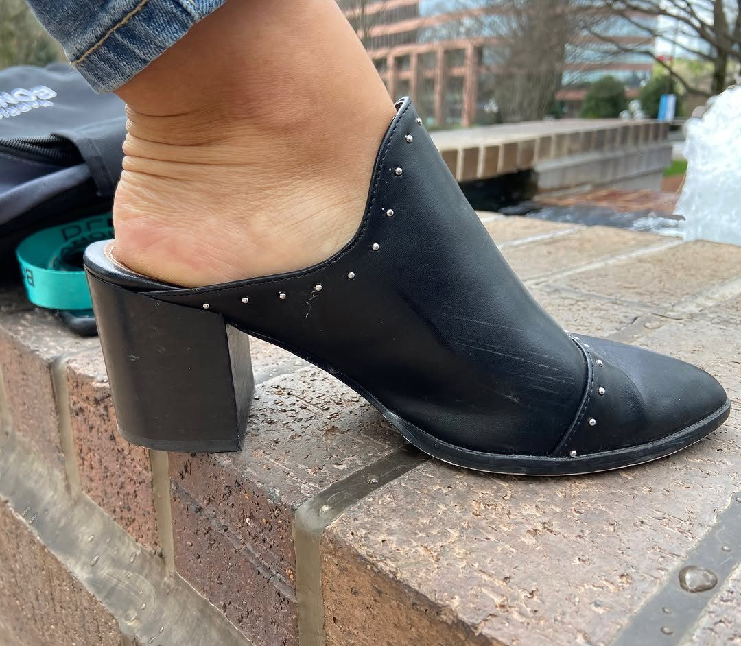 Black leather heeled clogs with silver studs worn on a brick ledge.