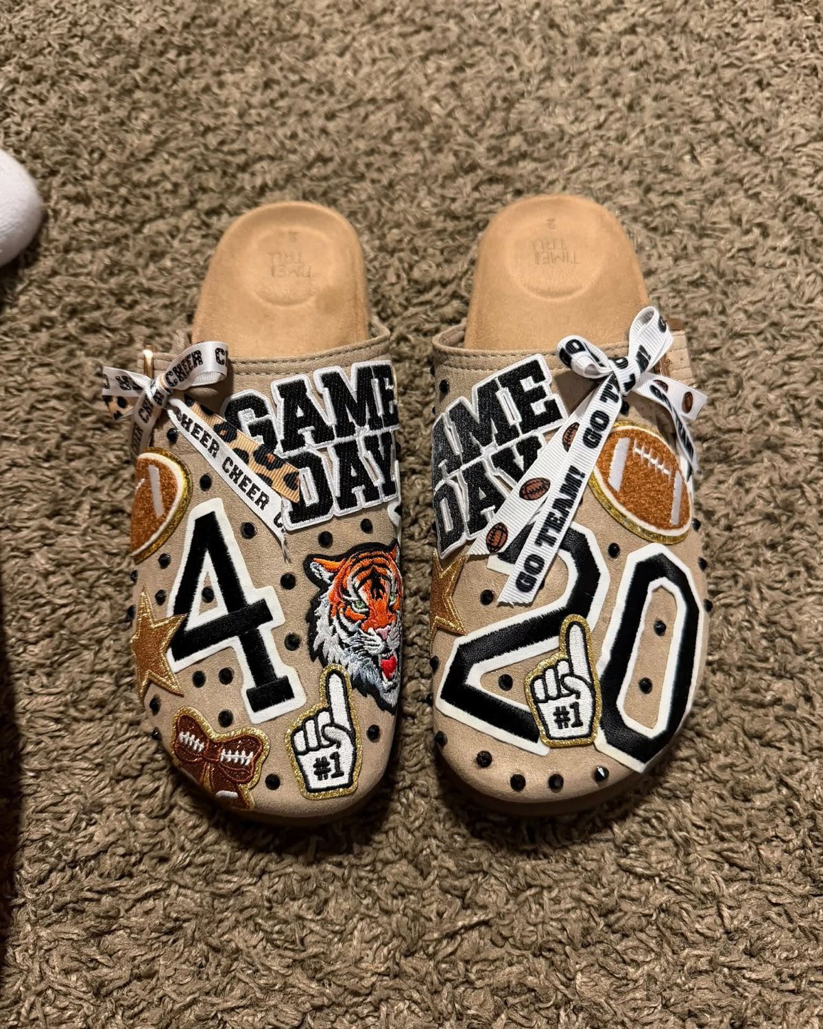 Tan clog shoes decorated with sports patches and ribbons on brown carpet.