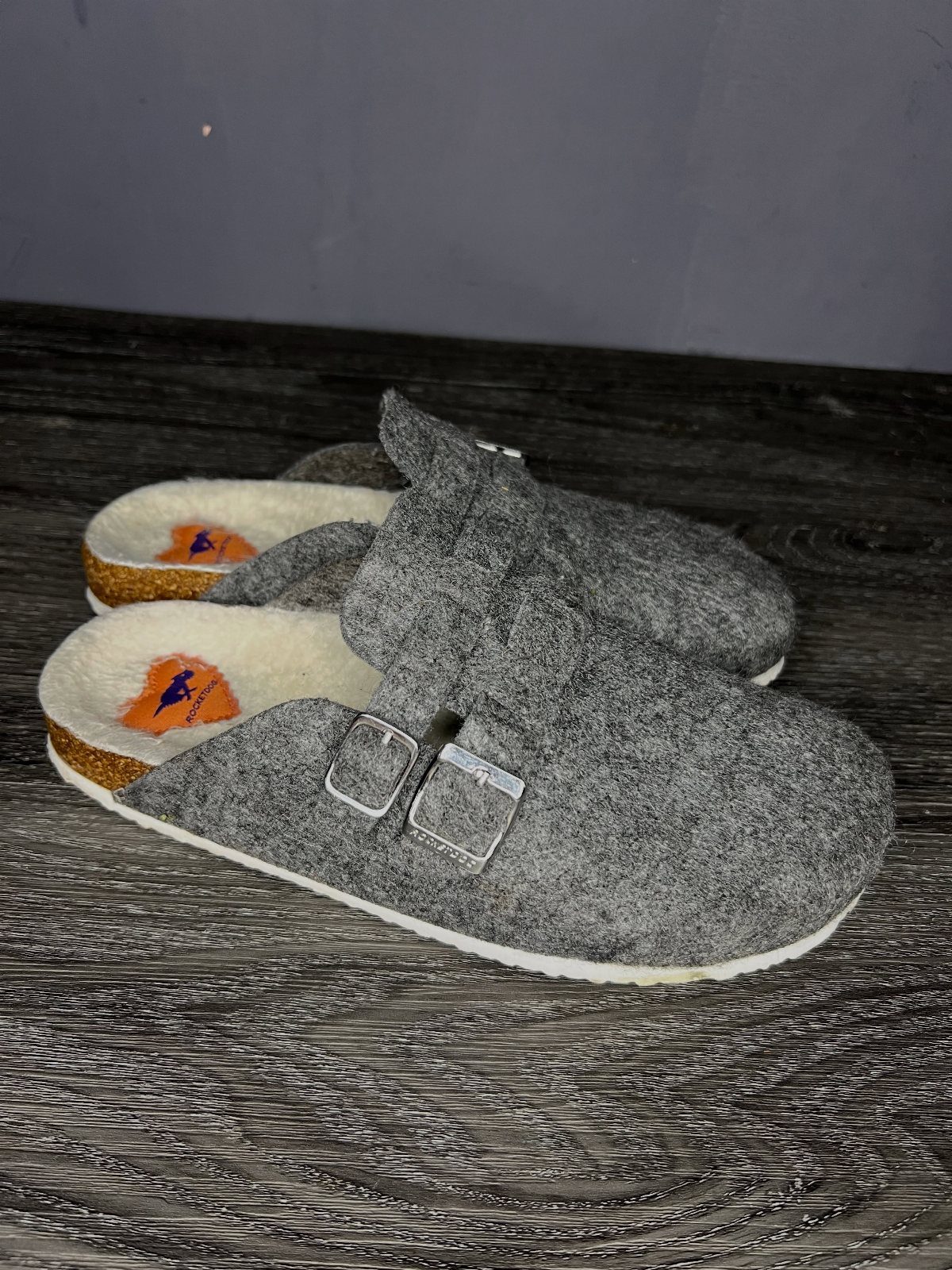 Grey felt clog mules with silver buckles and cork soles on dark wood.
