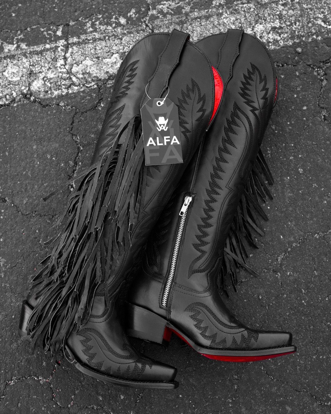 Black leather cowgirl boots with fringe and red soles on dark asphalt.