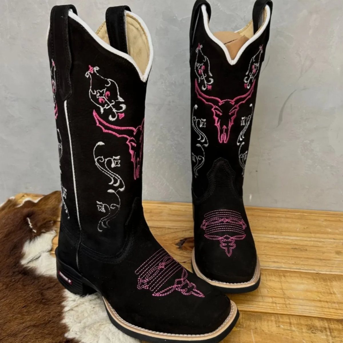 Black suede cowgirl boots with pink and white embroidery on a wooden surface.