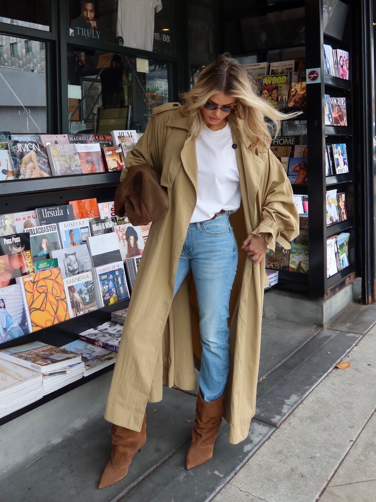 Woman wearing a tan trench coat, white shirt, and blue jeans by a magazine stand