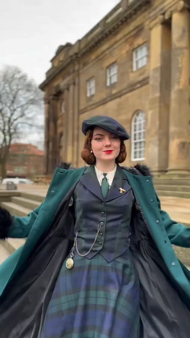 Woman in a green plaid skirt, vest, and beret standing before a stone building.