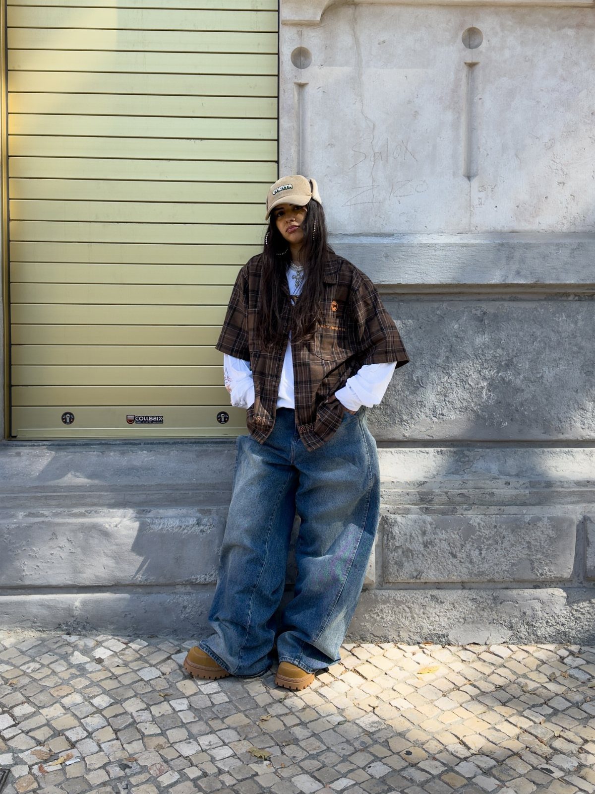 Woman wearing oversized plaid shirt, wide-leg blue jeans, and a tan baseball cap.