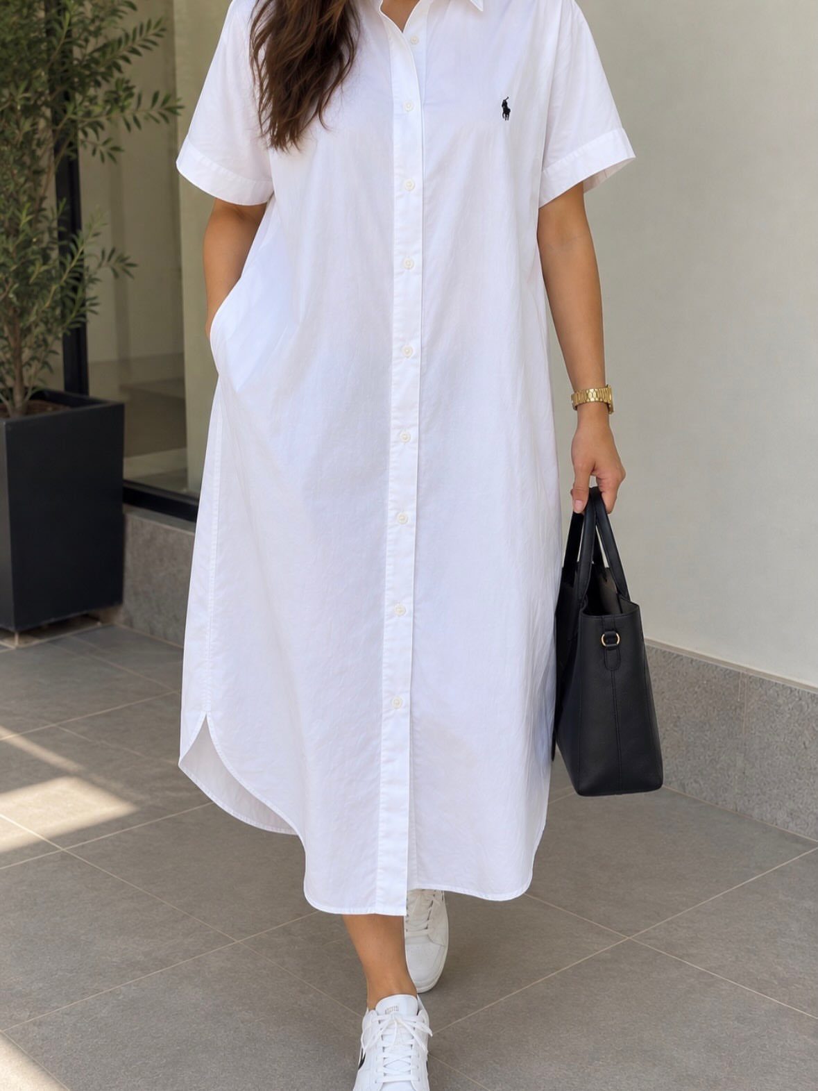 Woman wearing a white short-sleeved shirt dress with a black handbag and sneakers.
