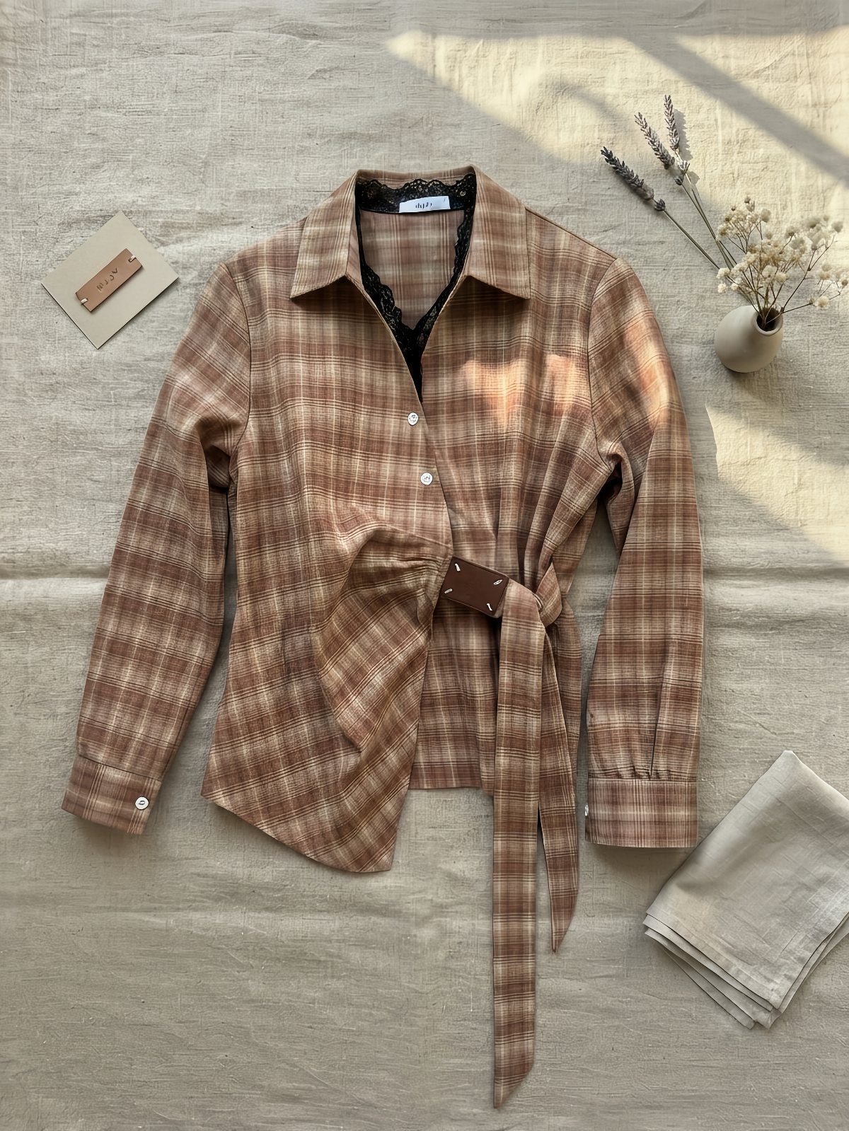Brown plaid button-down shirt with a tie belt on a linen background.