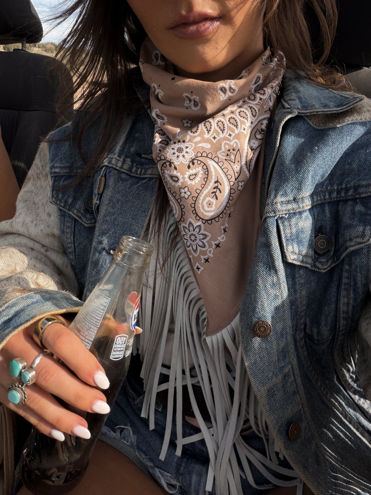 Person wearing a denim jacket, tan paisley bandana, and white fringed top.