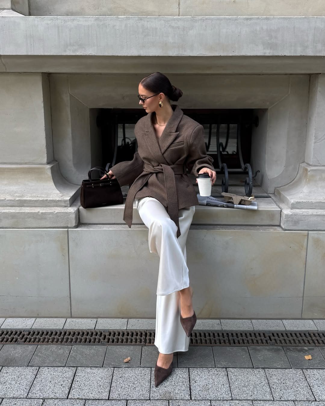 Woman in brown belted blazer and white trousers sitting on stone ledge