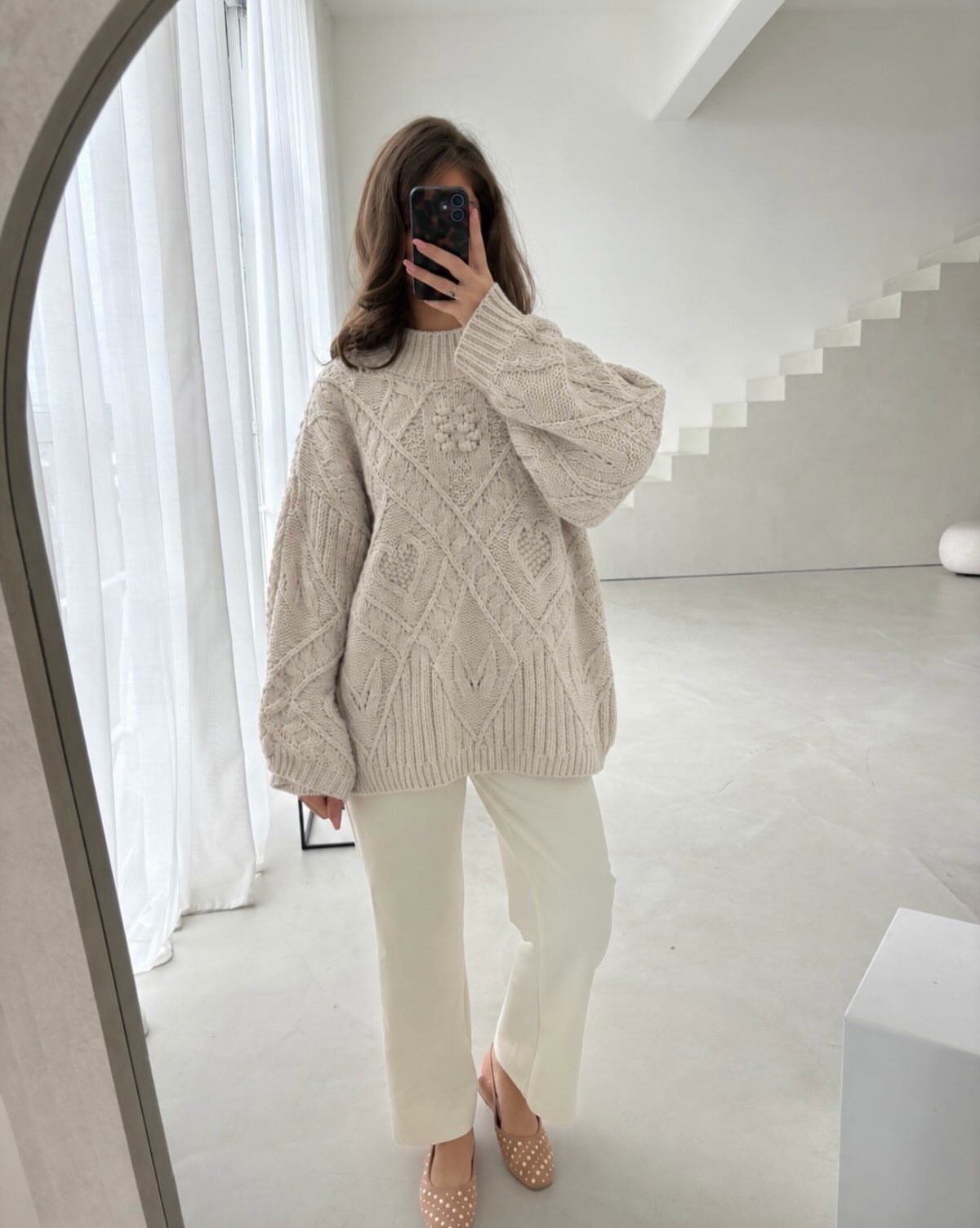 Woman in oversized beige cable-knit sweater and cream wide-leg trousers taking mirror selfie.