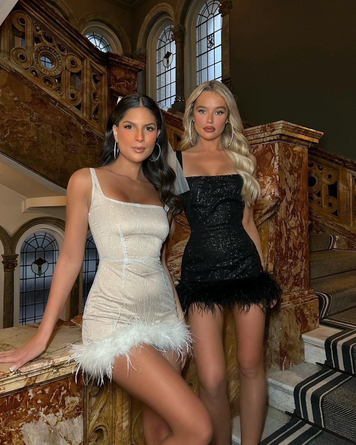 Two women in short shimmering white and black dresses with feather hems posing indoors.