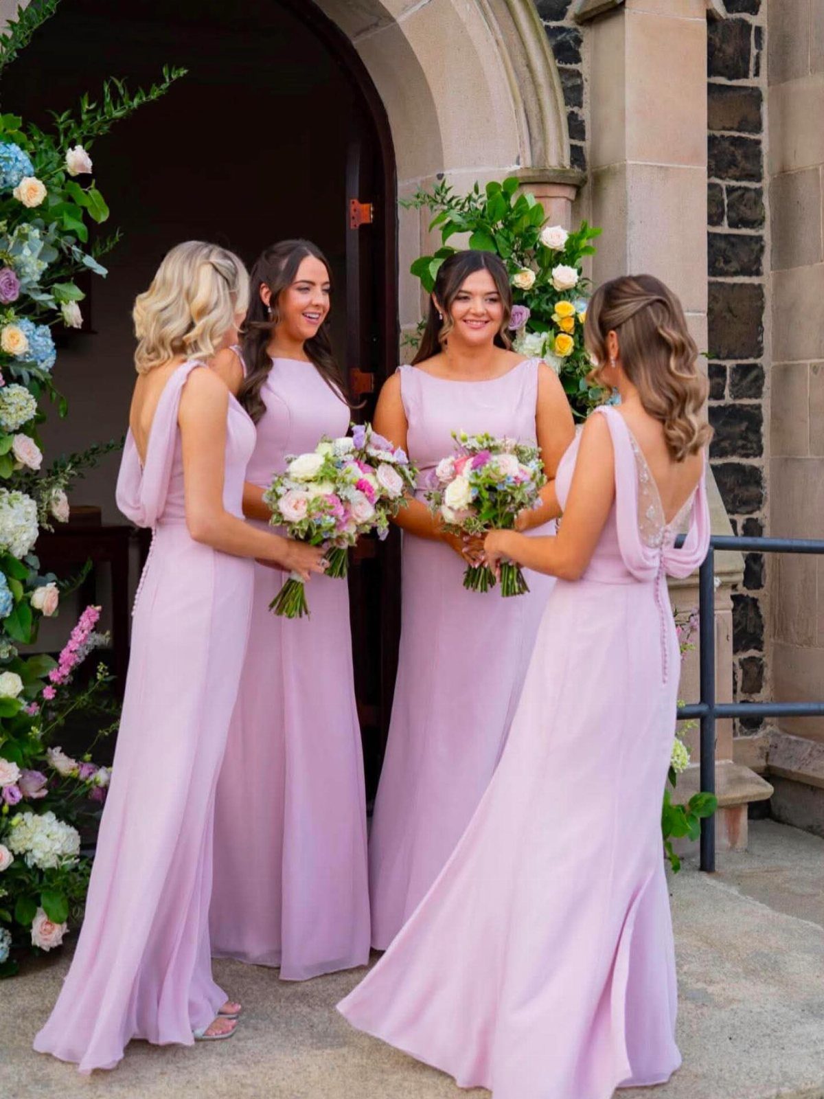 Four bridesmaids in light purple dresses holding floral bouquets outside a stone building.