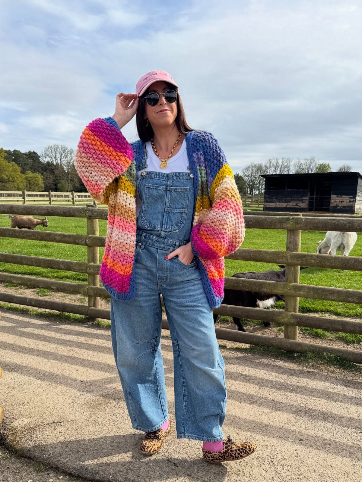 Woman in colorful cardigan and denim overalls standing by a farm fence.
