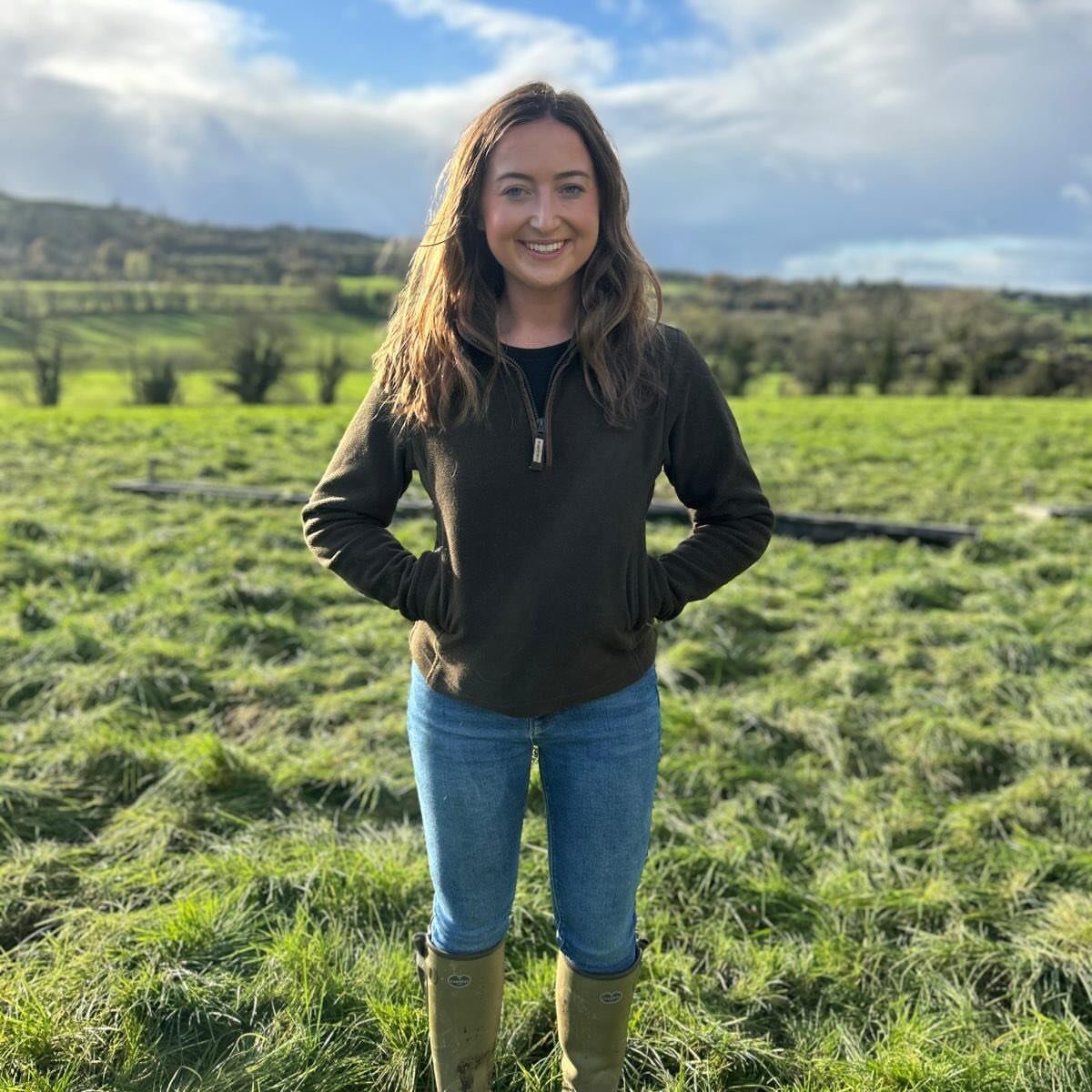 Woman in olive fleece and blue jeans standing in a green farm field