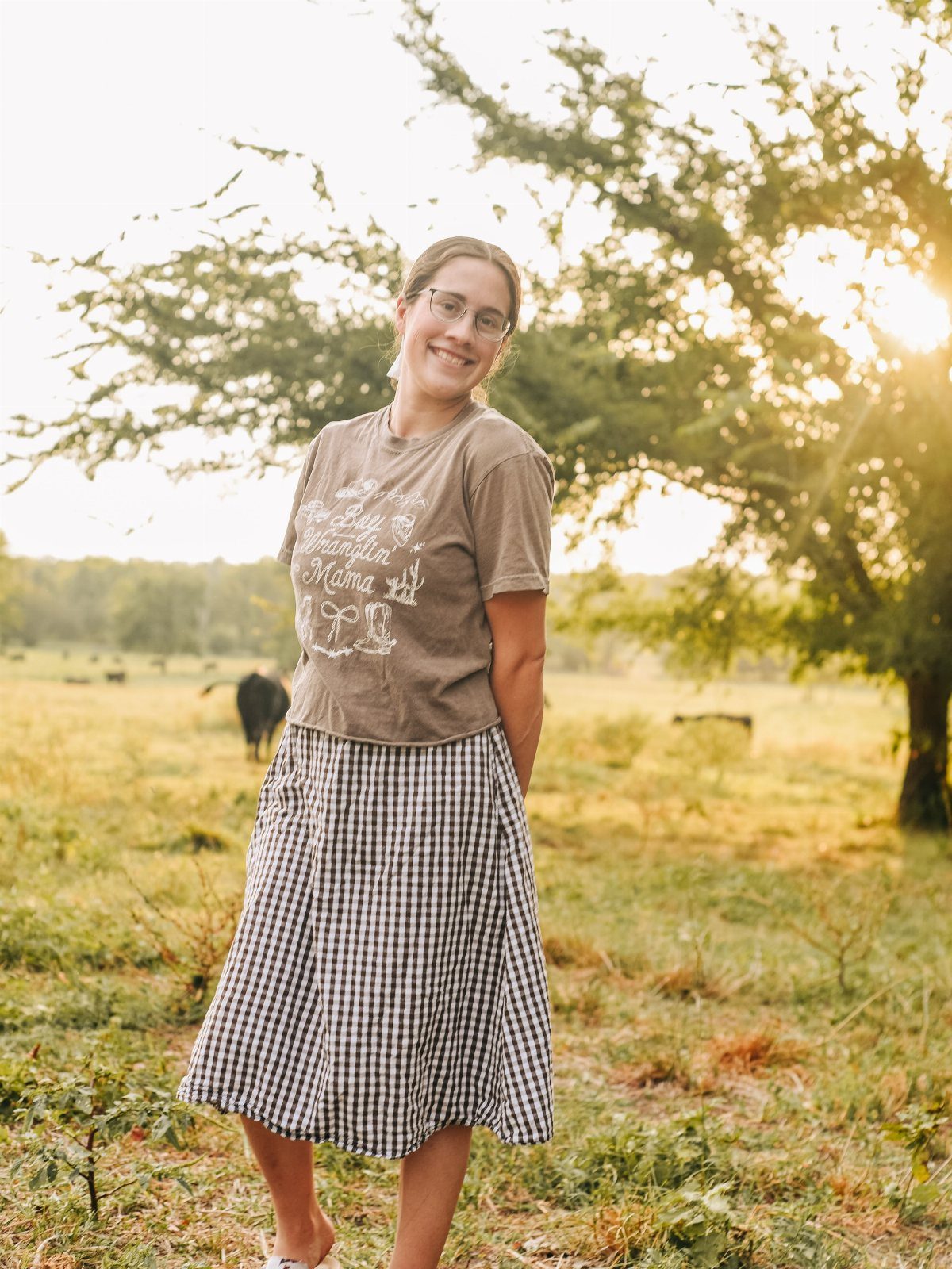 Woman in brown t-shirt and gingham skirt standing in a sunny farm field.