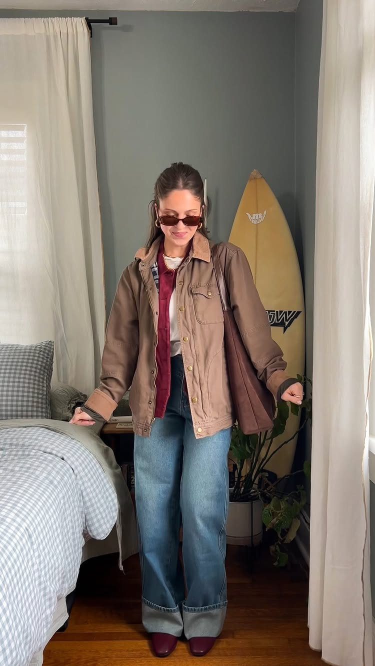 Woman wearing a brown jacket, red cardigan, and wide-leg jeans in a bedroom.