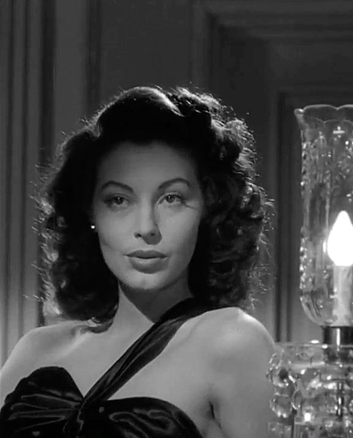 Femme Fatale 1940s Style: 5 Ideas from Film Noir 1 Woman with wavy hair wearing a dark satin gown next to a lamp.