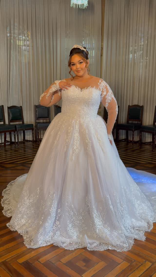Woman in a voluminous white lace wedding dress and tiara standing indoors.
